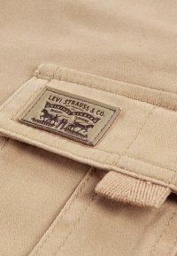 Μπεζ βαμβακερό ύφασμα με ραμμένη ετικέτα, που απεικονίζει γραφικό με άλογα και κείμενο, "Levi Strauss & Co. Original Riveted Quality Clothing."