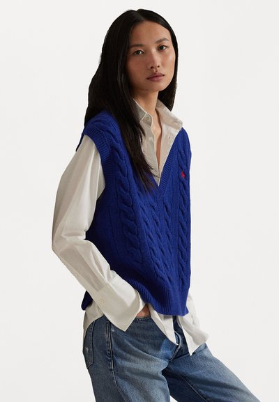 Blue cable-knit vest with V-neck, worn over a white button-up shirt. Loose fit with pronounced cable pattern. Paired with denim jeans.