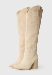 Bottes en daim beige montantes avec un bout pointu et un talon bloc. Présentent des coutures décoratives et un bord supérieur courbé pour plus de détails.