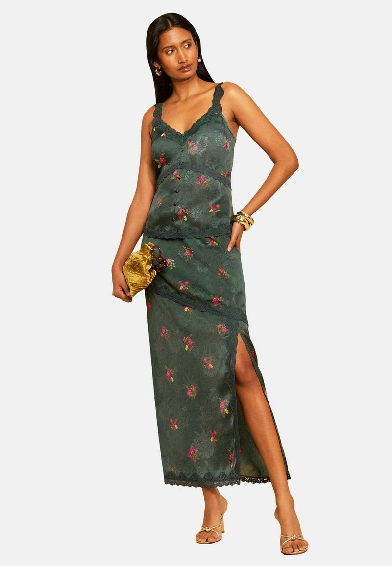Woman wearing a green floral sleeveless top and matching long skirt with a side slit, holding a gold pouch and wearing gold sandals and bracelets.