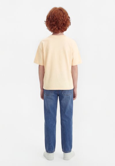 Niño con cabello rizado y rojo de espaldas, vestido con una camiseta amarillo claro, jeans azules y zapatillas blancas sobre un fondo blanco.
