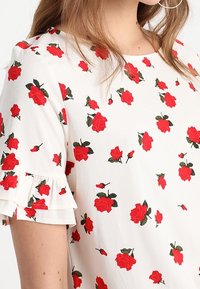Blusa blanca con mangas cortas rizadas, que presenta un patrón floral de rosas rojas y hojas verdes en toda la tela.