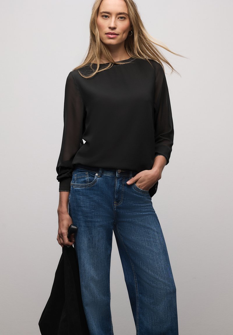 Blusa nera trasparente a maniche lunghe abbinata a jeans denim blu larghi. La blusa ha una scollatura tonda, e i jeans presentano tasche frontali.