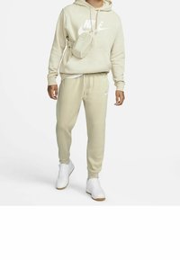 Béžová mikina Nike s bílým logem, odpovídající tepláky a malá béžová ledvinka. Bílé tenisky s gumovou podrážkou dokončují outfit.