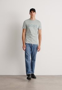 Hombre caminando hacia adelante, vistiendo una gorra negra, una camiseta gris clara con el texto "BOSS", jeans azules y zapatillas negras, frente a un fondo blanco liso.