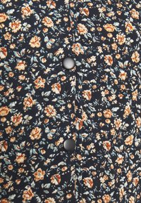 Veste matelassée en tissu bleu marine foncé avec un motif floral orange, crème et vert. Dotée de boutons noirs et d'un motif de couture en losange.