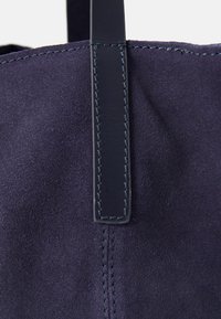 A navy blue suede tote bag features a smooth leather handle, reinforced stitching, and a soft, textural finish.
