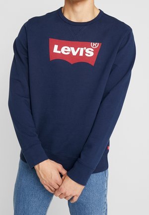 Personne portant un sweat-shirt Levi's bleu marine avec un logo rouge et blanc sur la poitrine et un jean bleu clair, les mains jointes devant.