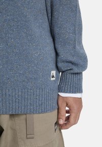 Blauer Strickpullover mit gerippten Bündchen, der mehrfarbige Sprenkel und ein Logotag am unteren Saum aufweist. Weißes Unterhemd ist an den Bündchen sichtbar.