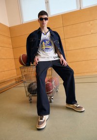 Džínová bunda a kalhoty v tmavě modré, bílý basketbalový dres s logem, hnědé tenisky, stojící vedle nákupního vozíku s basketbalovými míči.