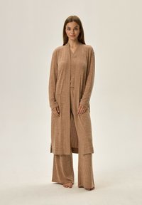 Jasnobrązowy zestaw odzieży domowej składający się z długiego kardiganu z kieszeniami, bluzki z dekoltem w serek oraz luźnych spodni wykonanych z miękkiego, teksturowanego materiału.