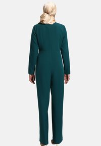 Mono de manga larga en verde azulado oscuro, con un cuerpo ajustado, piernas rectas y una tela suave y texturada. Acompañado de una bufanda dorada clara.