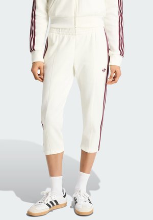 Persoon draagt witte cropped trainingsbroek met bordeauxrode zijstrepen en bijpassend jasje, witte sokken en witte sneakers met zwarte strepen.