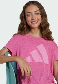 Roza heather majica s preklopljenim rukavima s velikim bijelim logotipom. Tkanina izgleda mekano s opuštenim krojem. Pozadina je neutralna.