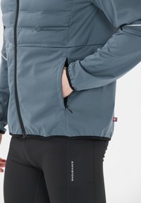 Person iført en blå lynlåsjakke med hånden i sidelommen og sorte leggings med teksten "Endurance" på låret.