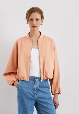 Veste bomber de couleur pêche avec un col montant, fermeture éclair et poignets élastiques. Portée par-dessus une chemise blanche et un jean bleu clair.