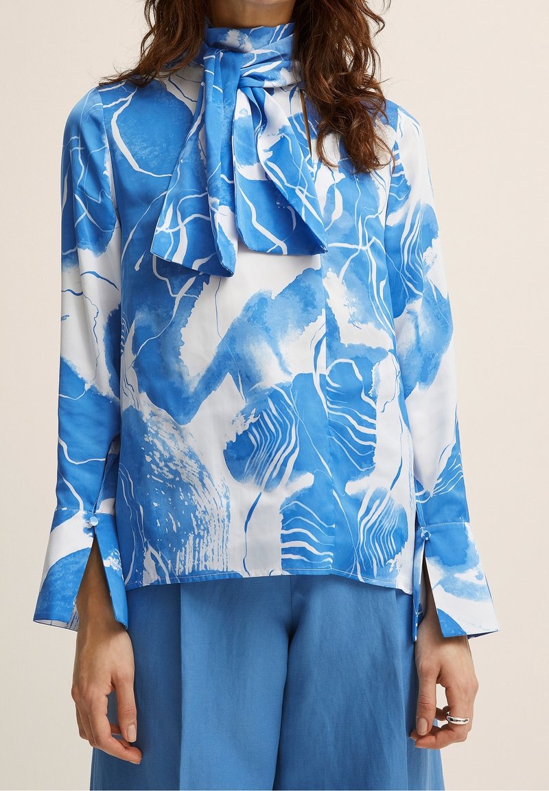 Blouse bleue et blanche avec motifs abstraits, col montant, détail lien, manches longues, fentes latérales, en tissu léger.