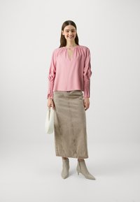 Blusa rosa con scollatura arricciata e maniche lunghe, abbinata a una gonna di jeans beige. La modella tiene una borsa a tracolla bianca, indossando stivali alla caviglia beige.