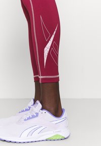 Leggings capri color bordeaux con grafiche astratte bianche, abbinati a scarpe da corsa viola chiaro e bianche con dettagli verdi e in rete.