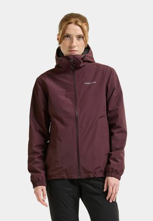 Vrouw met rood haar die een kastanjebruine waterdichte jas met capuchon en een zwarte broek draagt, staand tegen een effen lichte achtergrond.