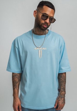 Mann mit Tattoos trägt blaues Dada T-Shirt, Sonnenbrille mit Kette und silberne Kettenhalskette vor einfachem grauem Hintergrund.