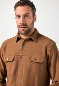 Brun struktureret skjorte med knaplukning foran, to brystlommer og spidse kraver. Har mørke knapper og en skræddersyet pasform.