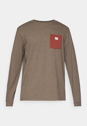Chemise à manches longues en coton brun foncé avec une poche rouge sur le côté gauche de la poitrine, arborant une petite étiquette de logo au-dessus de la poche.