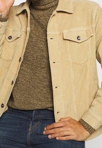 Homme portant une veste en velours côtelé beige sur un pull en tricot olive et un jean bleu, la main reposant sur la hanche.