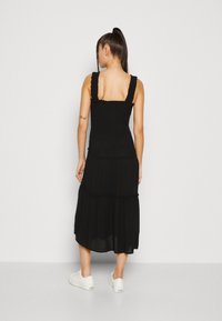 Robe noire midi avec des bretelles froncées, un corsage froncé et un détail de jupe à volants. Le mannequin porte des baskets blanches. Tissu lisse et léger.