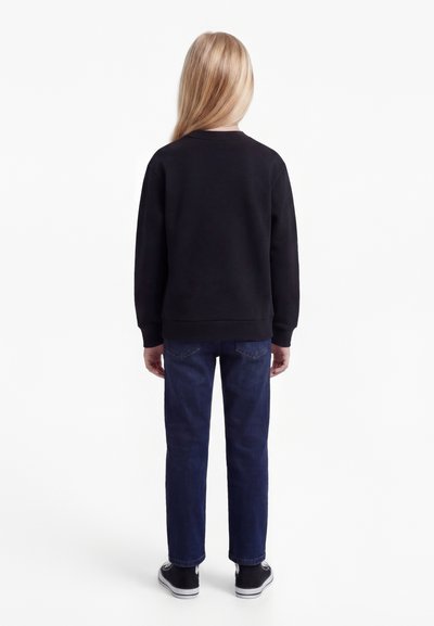 Copil cu păr lung și blond, purtând un hanorac negru, blugi albaștri și încălțăminte sport neagră, stând cu fața spre spate pe un fundal alb.