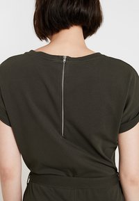 Persona con cabello corto y oscuro que lleva una camiseta verde oscuro con una cremallera plateada visible que recorre el centro de la espalda.