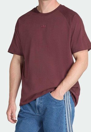 Mand iført en mørkerød Adidas t-shirt og blå jeans med hvide striber, hånd i lommen, foran en ensfarvet baggrund.