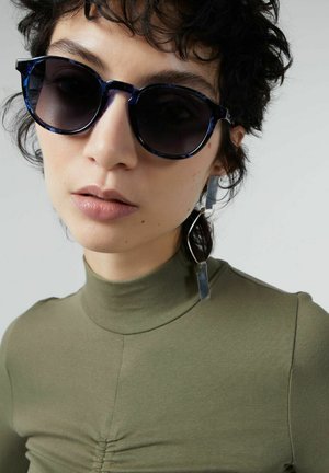 Round sunglasses with a navy tortoiseshell frame, dark lenses, worn with an olive green fitted top featuring a high neckline and subtle gathers.
