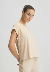 Top beige a maniche corte in tessuto morbido con vestibilità ampia, scollo rotondo e sottili spacchi laterali per un comfort e stile aggiuntivi.