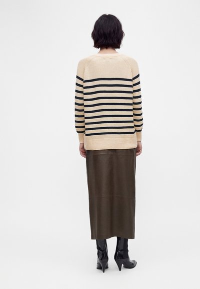 Personne aux cheveux courts et foncés portant un pull beige et noir rayé, une jupe midi en cuir marron et des bottes noires à talons, debout de dos.