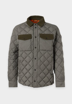 Jeune homme portant une veste matelassée vert olive, un pantalon large marron, des baskets beiges, debout devant un fond clair uni.