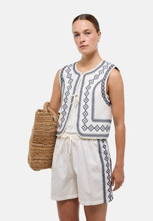 Femme portant un gilet sans manches blanc et un short assorti avec des motifs géométriques noirs, tenant un grand sac en paille tressée.