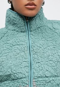 Miętowa, teksturowana kurtka polarowa z wysokim kołnierzem i zamkiem z przodu, charakteryzująca się miękką, puszystą powierzchnią oraz kontrastującym wykończeniem wokół zamka.
