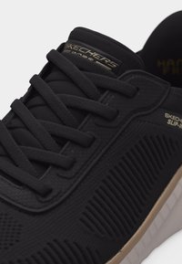 Κοντινό πλάνο μαύρου αθλητικού παπουτσιού Skechers με υφή υφάσματος, μαύρα κορδόνια και αντίθετη μπεζ σόλα.