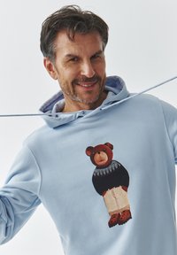 Sudadera azul claro con una ilustración de un oso con un suéter negro y pantalones beiges, decorada con acentos de patrón y botas naranjas.