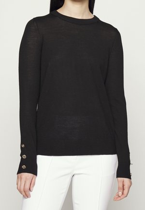 Femme portant un pull noir à manches longues avec trois boutons dorés sur chaque poignet et un pantalon blanc, montrée du cou jusqu'aux hanches.