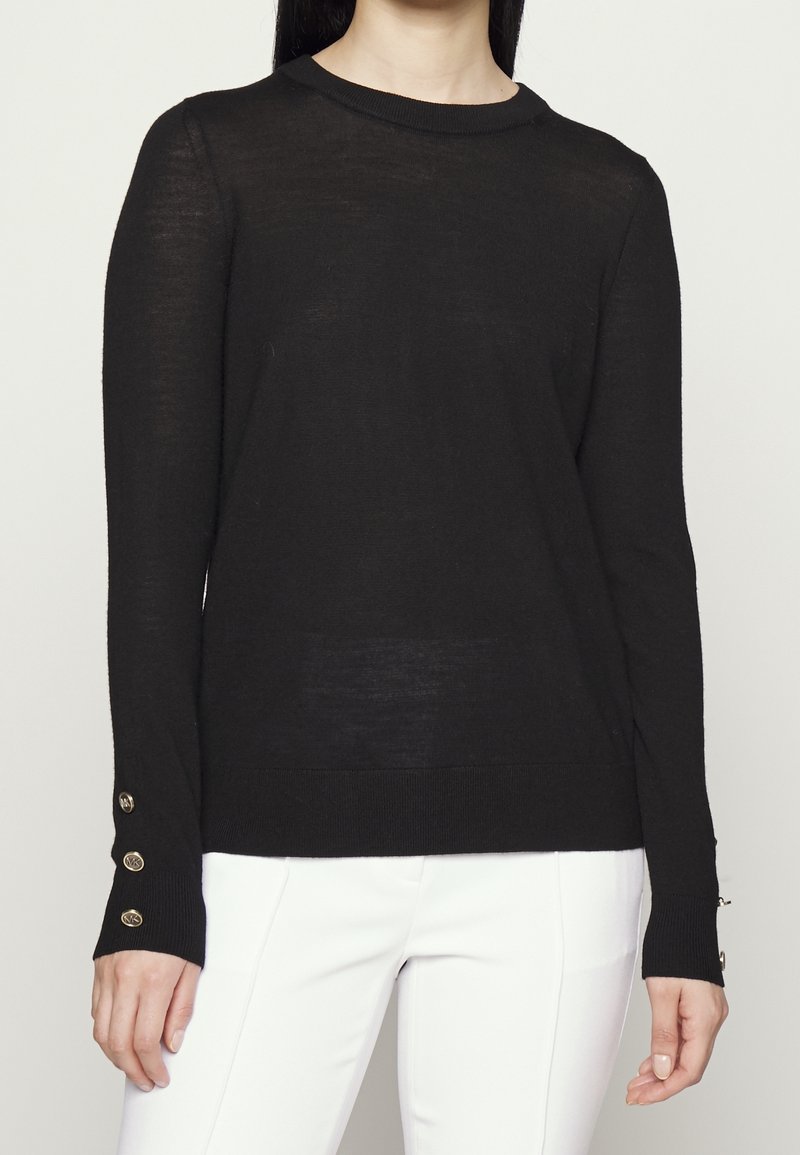Femme portant un pull noir à manches longues avec trois boutons dorés sur chaque poignet et un pantalon blanc, montrée du cou jusqu'aux hanches.