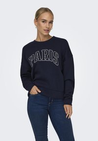 Jūras zila džemperis ar baltiem burtiem "PARIS", apaļš kakls, brīvā piegriezumā un ribotām piedurknēm. Uzvilkts kopā ar džinsa biksēm, modelis rokas ielicis kabatās.