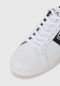 Adidași din piele albă, cu vârf rotund, șireturi albe și un accent negru cu branding pe lateral. Talpă din cauciuc texturat pentru aderență.