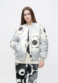 Jasnoszara kurtka bombowiec z dużym czarnym kwiatowym wzorem, ściągaczami przy rękawach i zamkiem błyskawicznym. Nosi się ją na kremowym swetrze i spodniach w kwiatowy wzór.