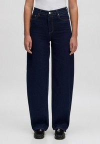 Donkerblauwe wijdvallende jeans van denim, met vijf zakken, een sluiting met een knop aan de voorkant en subtiele stikseldetails. Gecombineerd met zwarte schoenen.