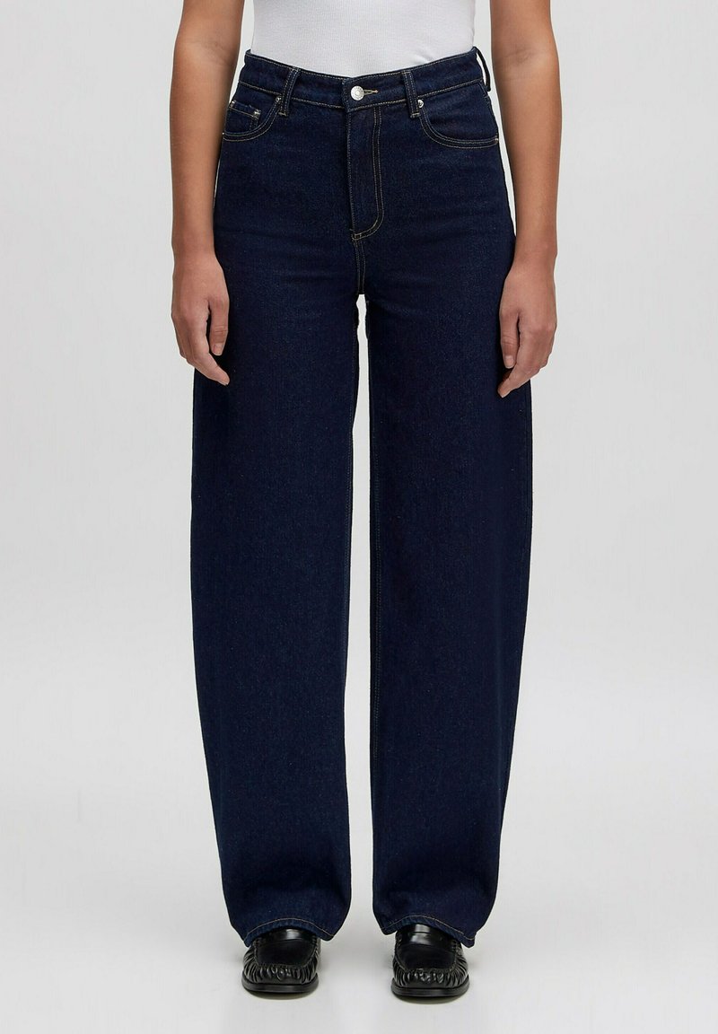 Donkerblauwe wijdvallende jeans van denim, met vijf zakken, een sluiting met een knop aan de voorkant en subtiele stikseldetails. Gecombineerd met zwarte schoenen.