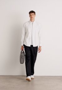Witte blouse, zwarte broek, witte sneakers en grijze tas. Model loopt op een lichte ondergrond met een effen achtergrond. Casual, strakke look.