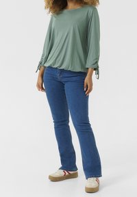 Světlý zelený svetr s dlouhými rukávy a nařasenými detaily na manžetách, v kombinaci s modrými džíny a béžovými teniskami s červenými pruhy. Hladká struktura látky.