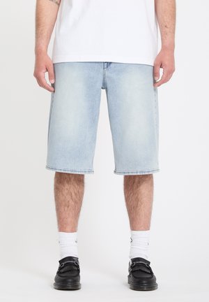 Homme portant un short en denim bleu clair, des chaussettes blanches, des chaussures noires et un t-shirt blanc, debout devant un fond blanc uni.